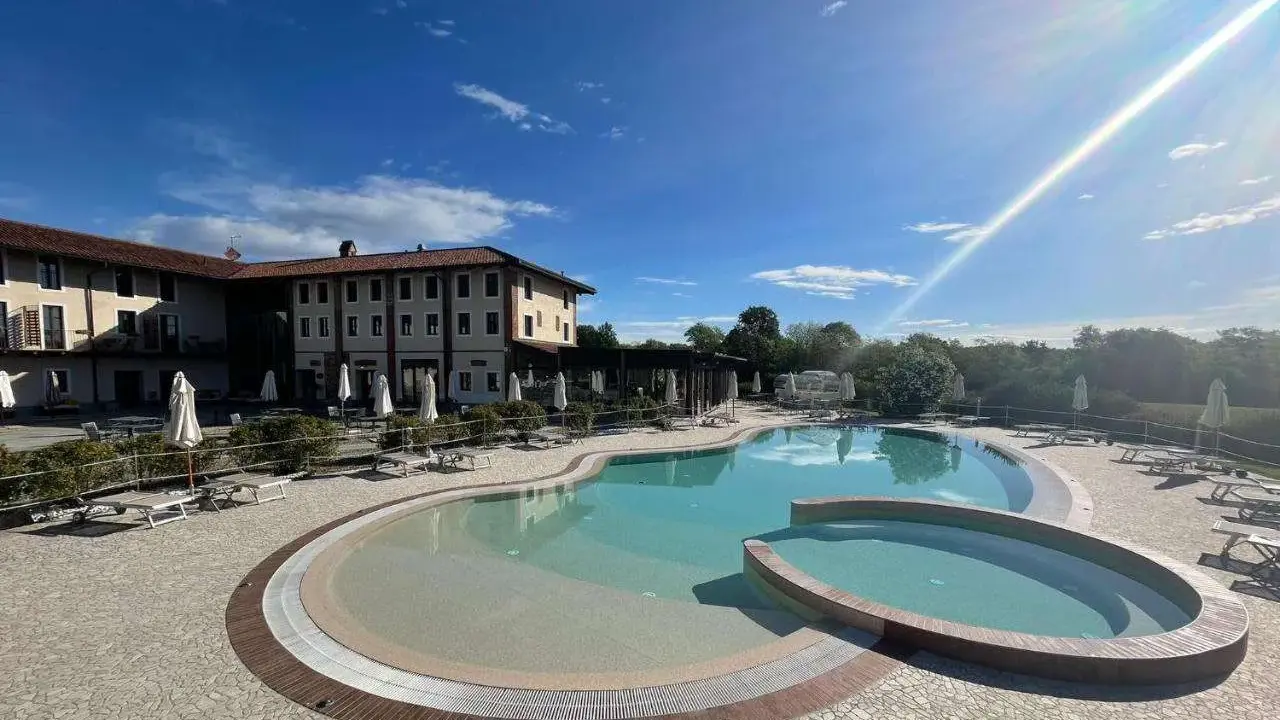 Piscina a forma di rene con idromassaggio annesso, circondata da lettini e ombrelloni, di fronte a un hotel in stile piemontese.