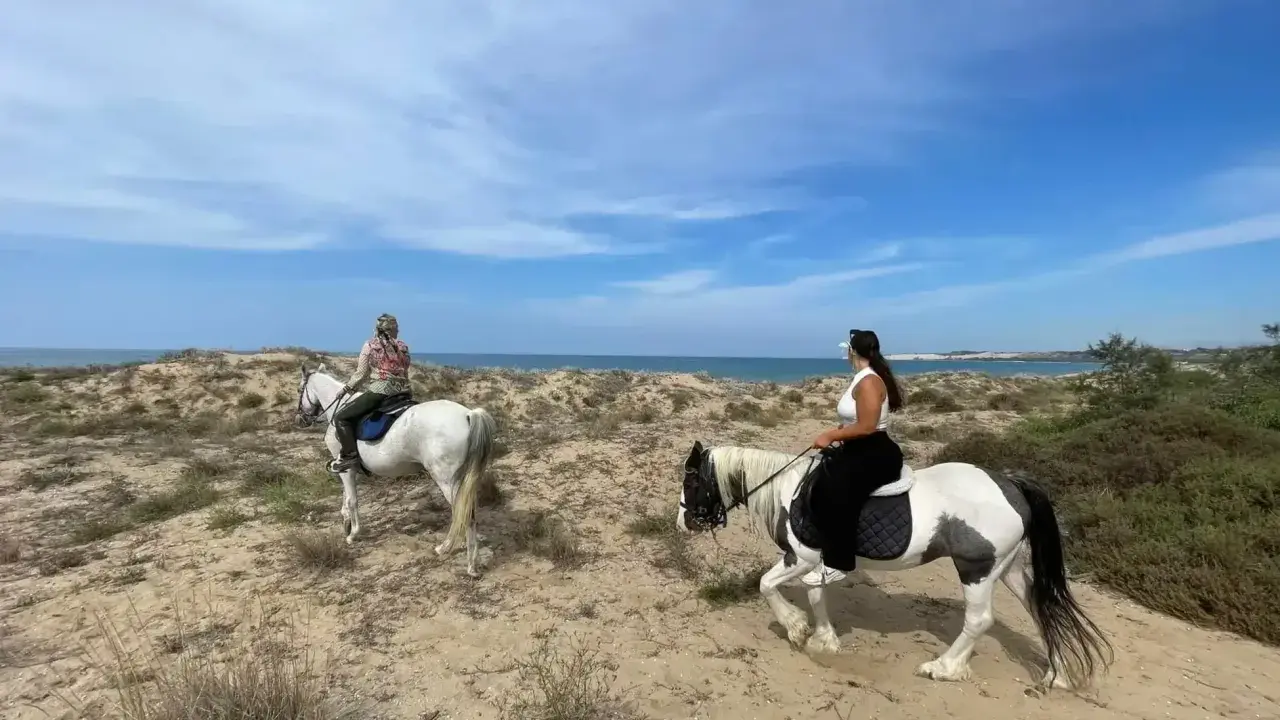 Due donne godono di una passeggiata a cavallo in Sicilia, tra dune sabbiose e il mare azzurro.