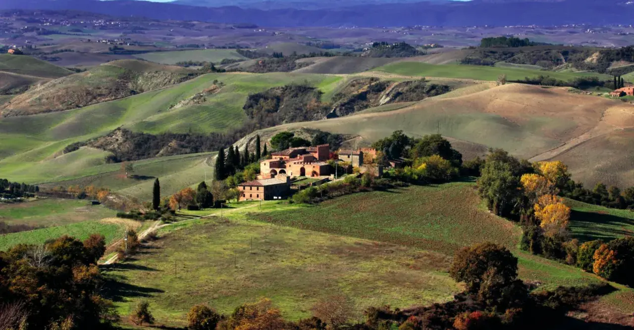 Colline ondulate e campi coltivati, un casale in mattoni rossi e cipressi svettanti, tipici delle **strade panoramiche toscana**.