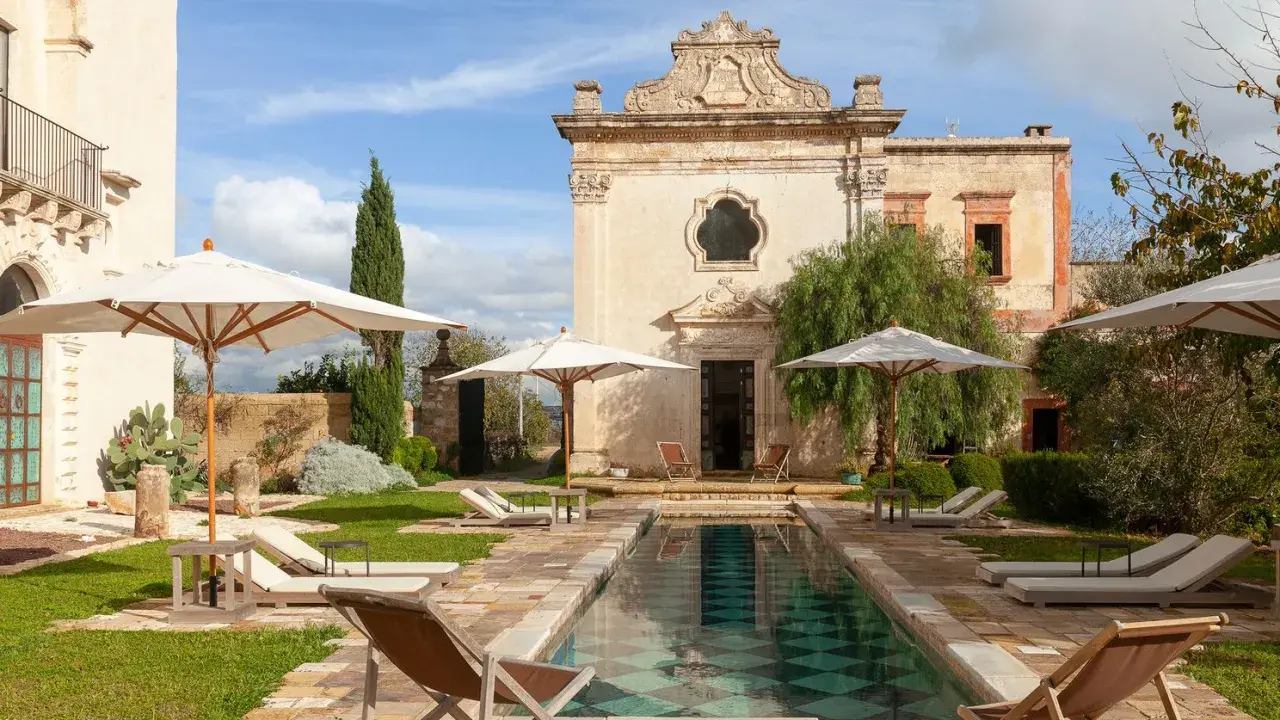 Piscina e lettini in una masseria per bambini in Puglia, con un antico edificio sullo sfondo.