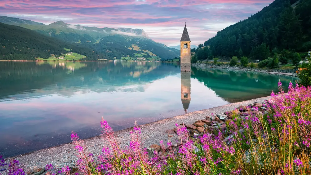 Torre sommersa in un lago alpino, con montagne verdi e cielo rosa all'alba. Un angolo di montagna Friuli Venezia Giulia incantevole.