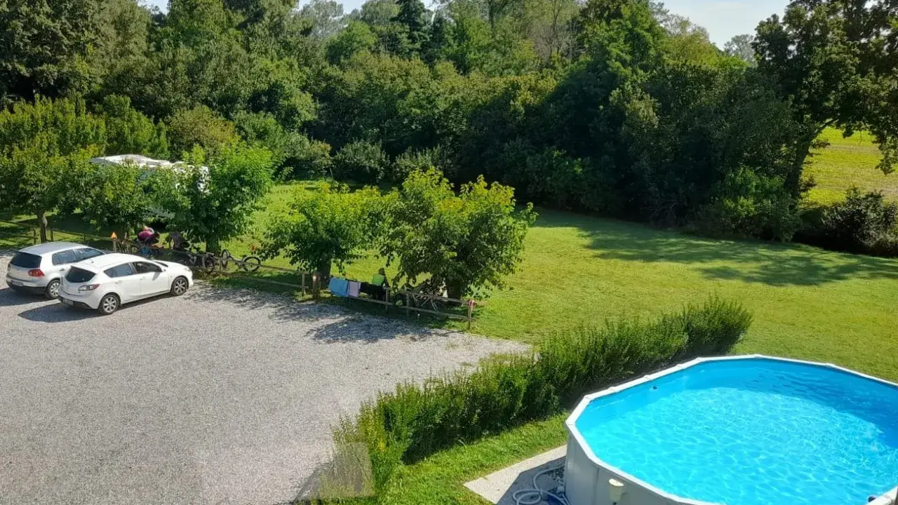 Un **agricampeggio con piscina** immerso nel verde. Auto parcheggiate, camper, biciclette e persone che si rilassano all'ombra degli alberi. La piscina azzurra invita a un tuffo rinfrescante.
