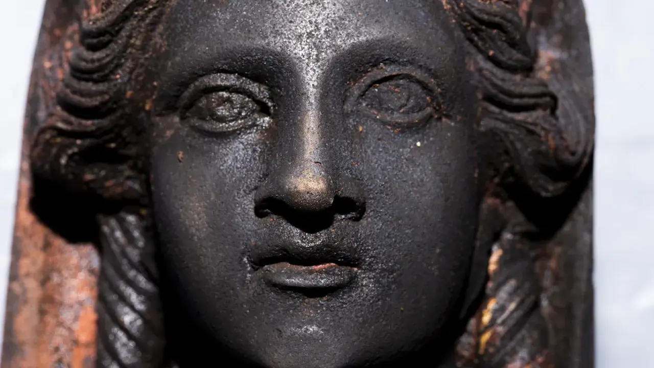 Volto di bronzo scuro, un dettaglio dei bronzi di San Casciano dei Bagni, con capelli a onde e trecce.