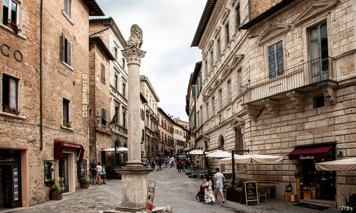 Un vicolo pittoresco tra i borghi vicino Montepulciano, con edifici storici, un leone scolpito su una colonna e tavolini all'aperto.