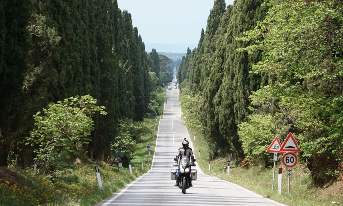 Un motociclista percorre un viale cipressi toscana, immerso nel verde e con il mare all'orizzonte.