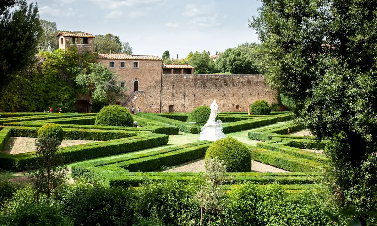 Giardino all'italiana con siepi geometriche, una statua bianca e un antico borgo toscano da visitare sullo sfondo.