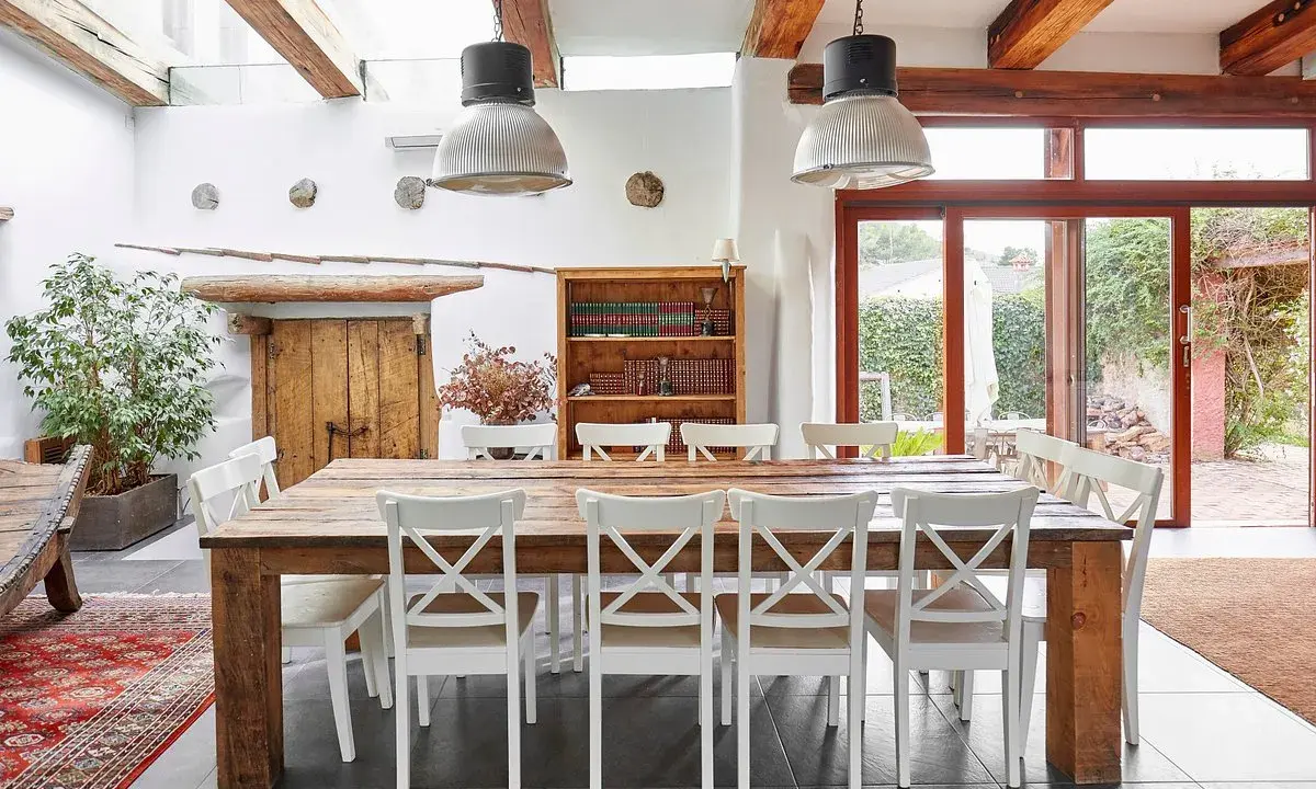 Un rustico tavolo in legno con sedie bianche, illuminato da lampade industriali, in una casa con travi a vista e un giardino che ricorda il Val di Cecina.