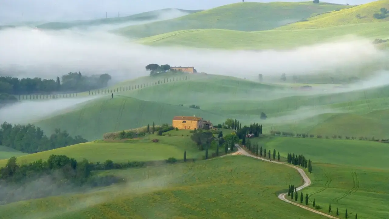 Dolci colline verdi avvolte dalla nebbia mattutina nel Val di Norcia, con casali in pietra e filari di cipressi.
