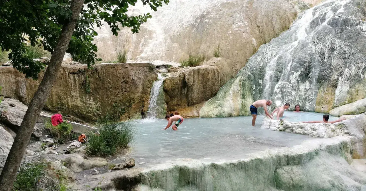 Persone si rilassano nelle piscine termali naturali di Castiglione d'Orcia, con cascate che scendono da rocce bianche.