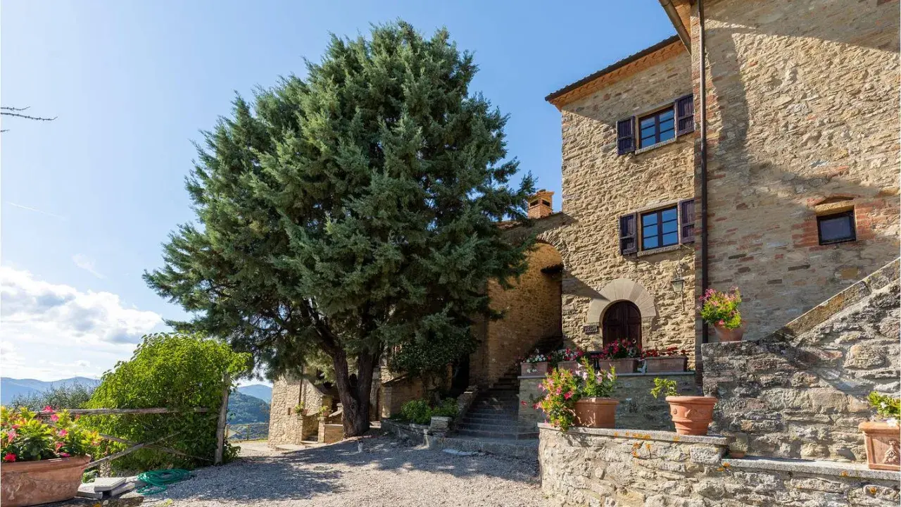 Un casale in pietra con un grande cipresso, tipico degli agriturismi del centro Italia, circondato da fiori in vaso e con un panorama collinare.
