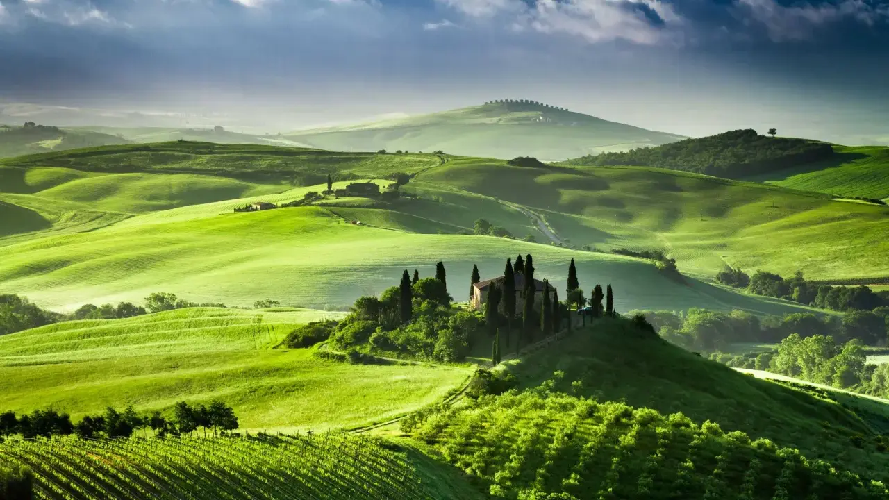 Colline verdi ondulate del Val d'Orcia, con una casa colonica e cipressi su una collina. Un paesaggio da sogno, quasi una mappa visiva della Toscana.