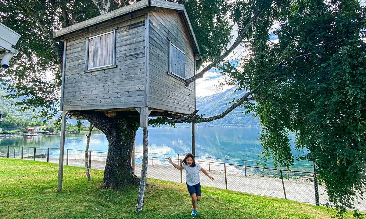Bambina corre verso una casa sull'albero in legno, con un lago e montagne sullo sfondo.