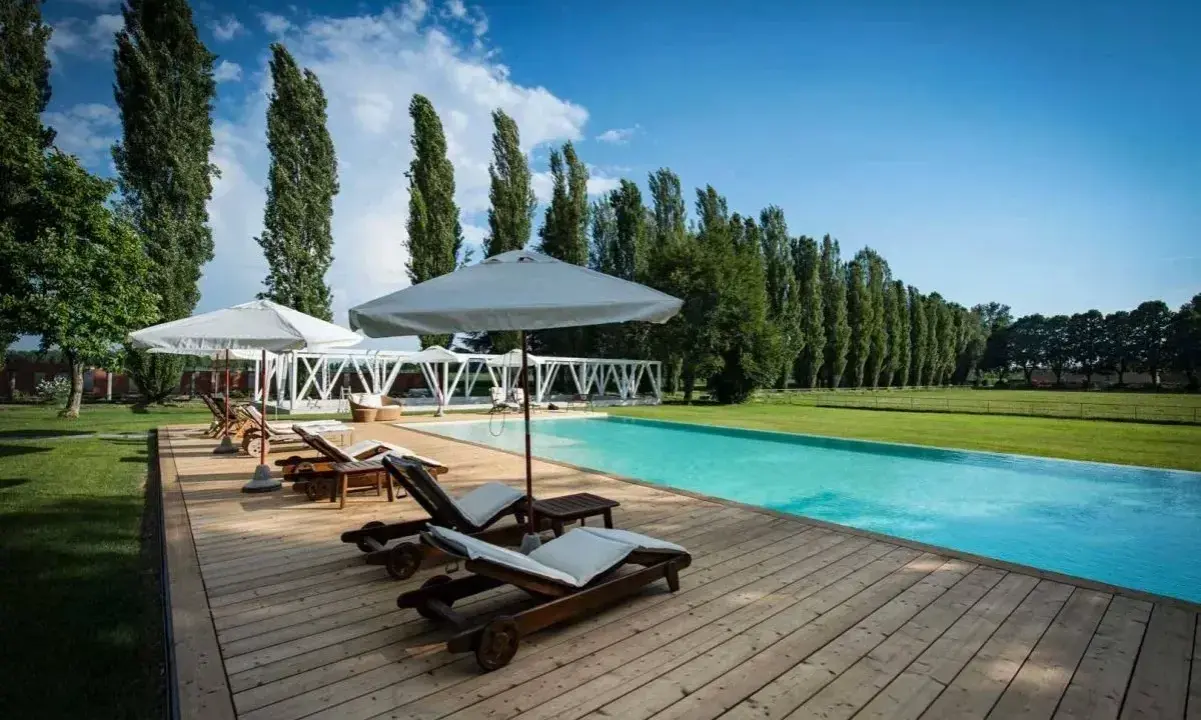 Una splendida piscina in campagna, con lettini prendisole in legno e ombrelloni bianchi, circondata da un prato verde e alti alberi.