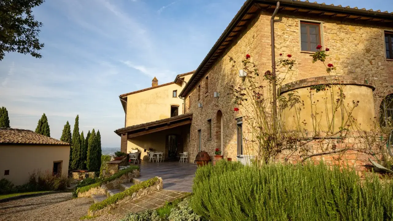Un casale in pietra con rose rampicanti, un pergolato con tavoli e sedie, e cipressi svettano nel paesaggio della **Toscana in inverno**.
