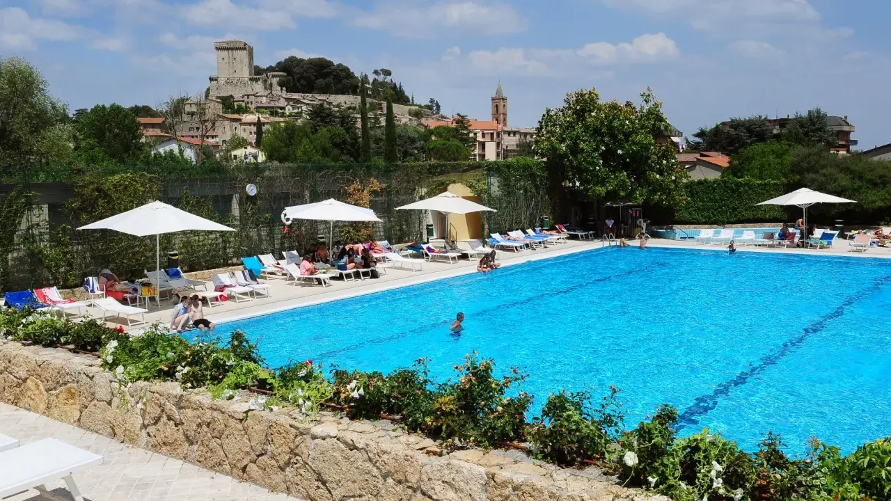 Piscina azzurra in un villaggio famiglie toscana, con castello medievale sullo sfondo e persone che si rilassano al sole.