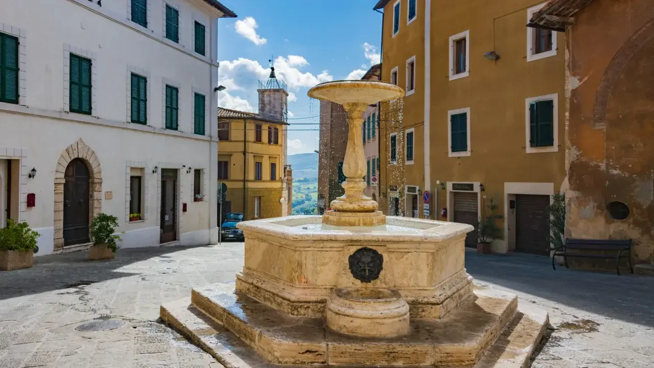 Fontana in pietra nella Valdichiana Senese, circondata da edifici storici con persiane verdi e un cielo azzurro con nuvole bianche.