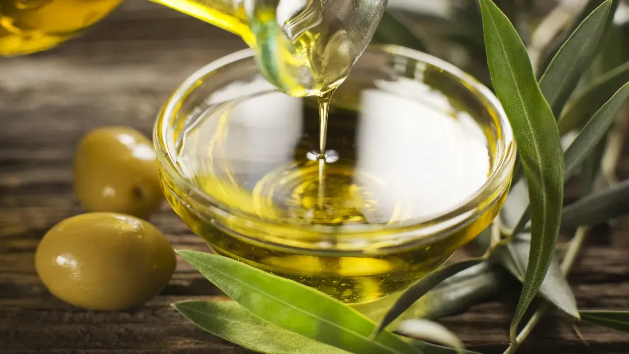 Olio di oliva non extravergine versato in una ciotola di vetro, con olive e foglie d'ulivo su fondo di legno.