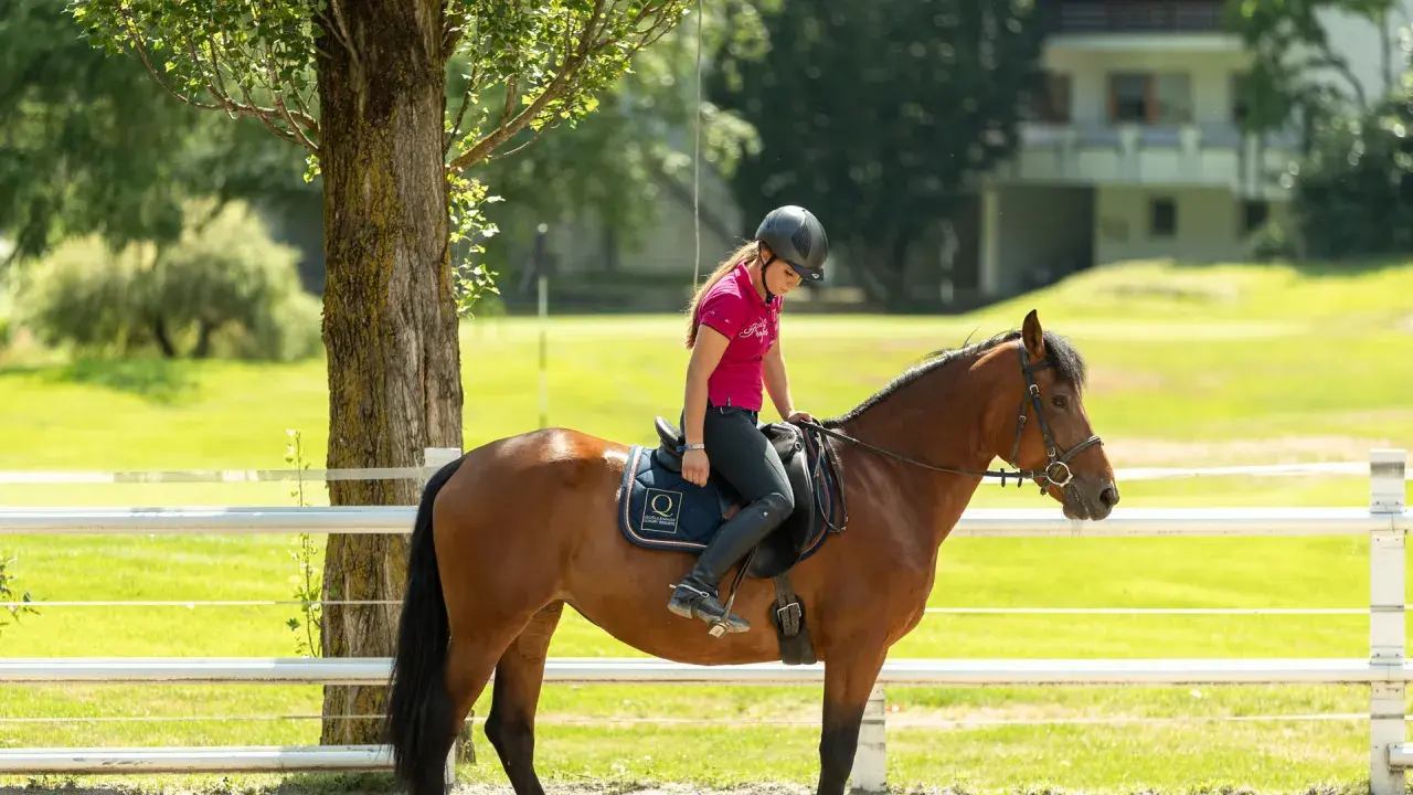 Ragazza in sella a un cavallo castano in un maneggio, con un edificio sullo sfondo. Un'esperienza indimenticabile in un hotel con maneggio.