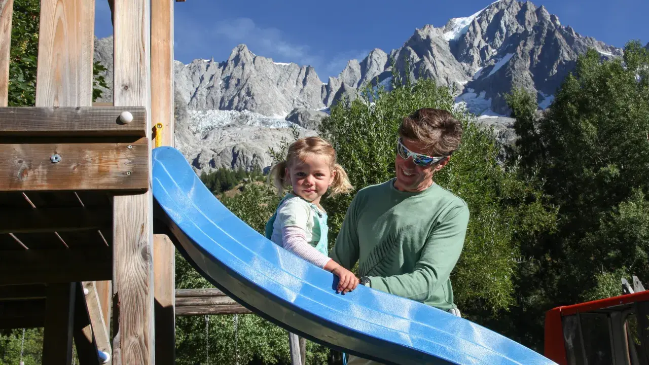 Un padre e una bambina si divertono su uno scivolo in un parco giochi con le maestose montagne della Valle d'Aosta sullo sfondo, un'estate indimenticabile.