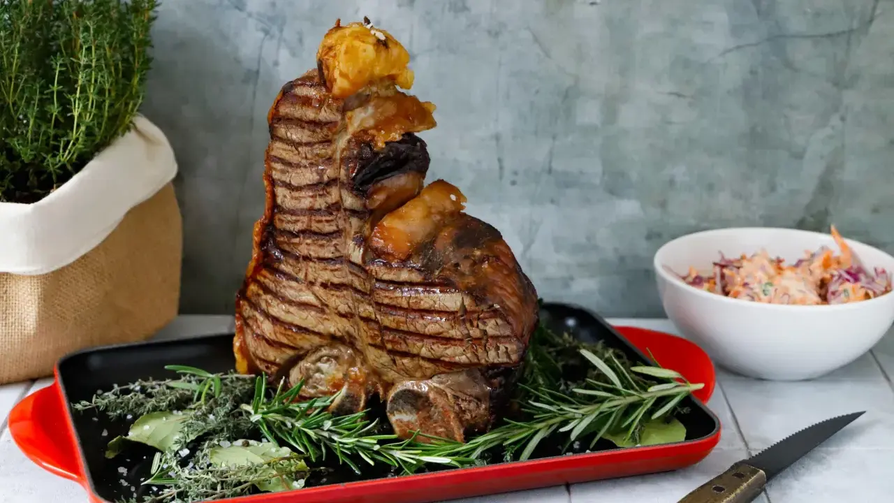 Una succulenta bistecca fiorentina, grigliata alla perfezione, adagiata su erbe aromatiche in una teglia rossa. Accanto, un'insalata fresca e un coltello.