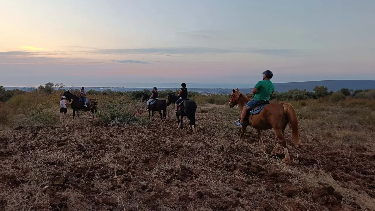 Un gruppo di persone fa una **passeggiata a cavallo in Puglia** al tramonto, godendosi il paesaggio collinare e il mare in lontananza.