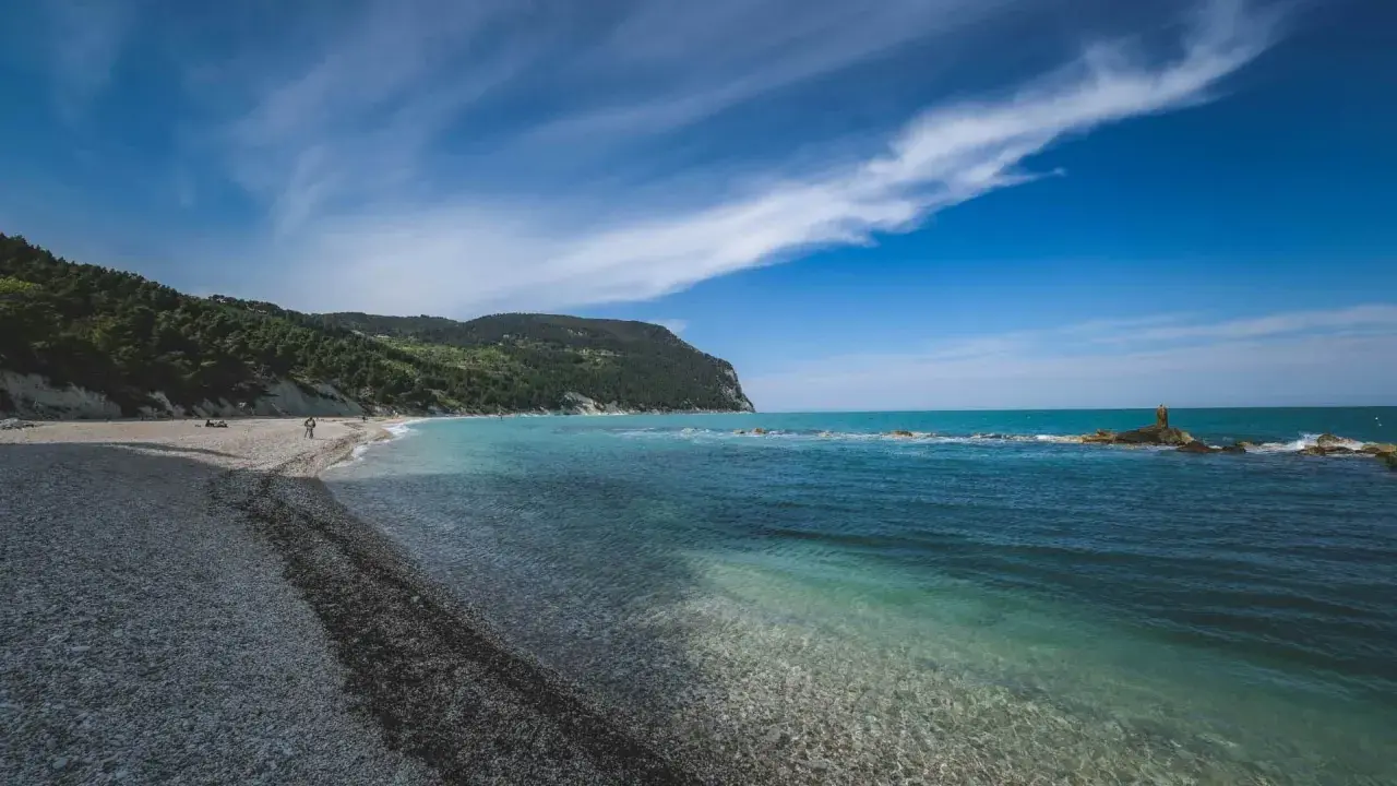 Spiaggia di ciottoli con mare cristallino e una collina verdeggiante, tipica del entroterra marchigiano.