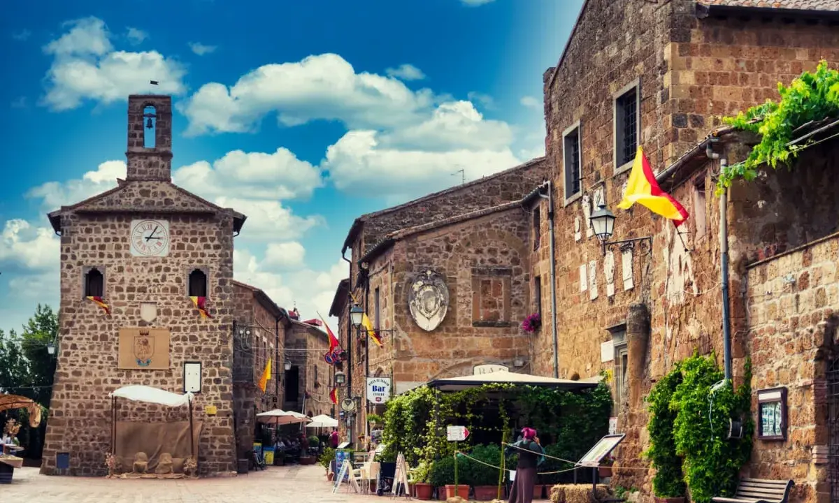 Un vicolo pittoresco a Sovana, Italia, con edifici in pietra, bandiere colorate e un cielo azzurro pieno di nuvole.