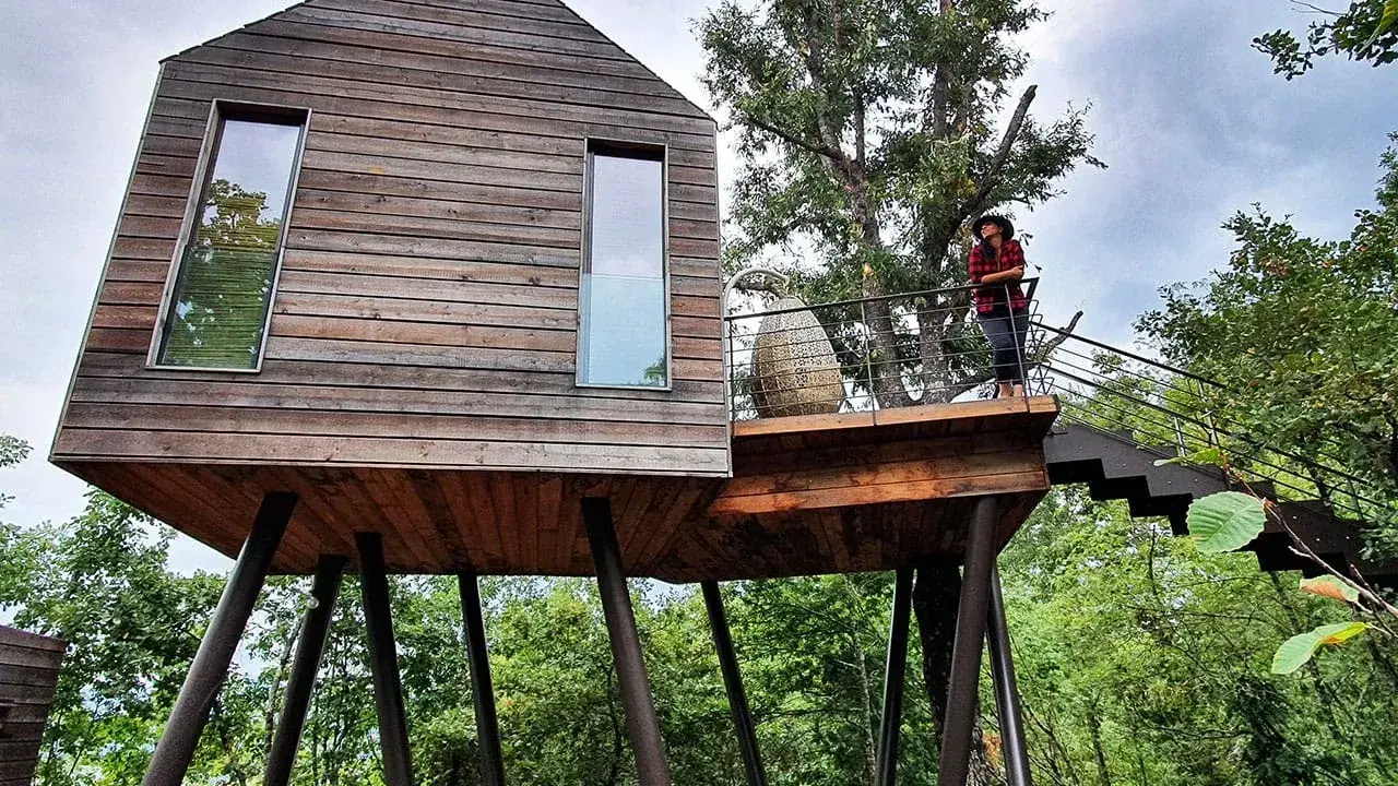 Un'esperienza unica per dormire nella natura toscana: una casa sull'albero in legno, con una persona che ammira il paesaggio dalla terrazza.