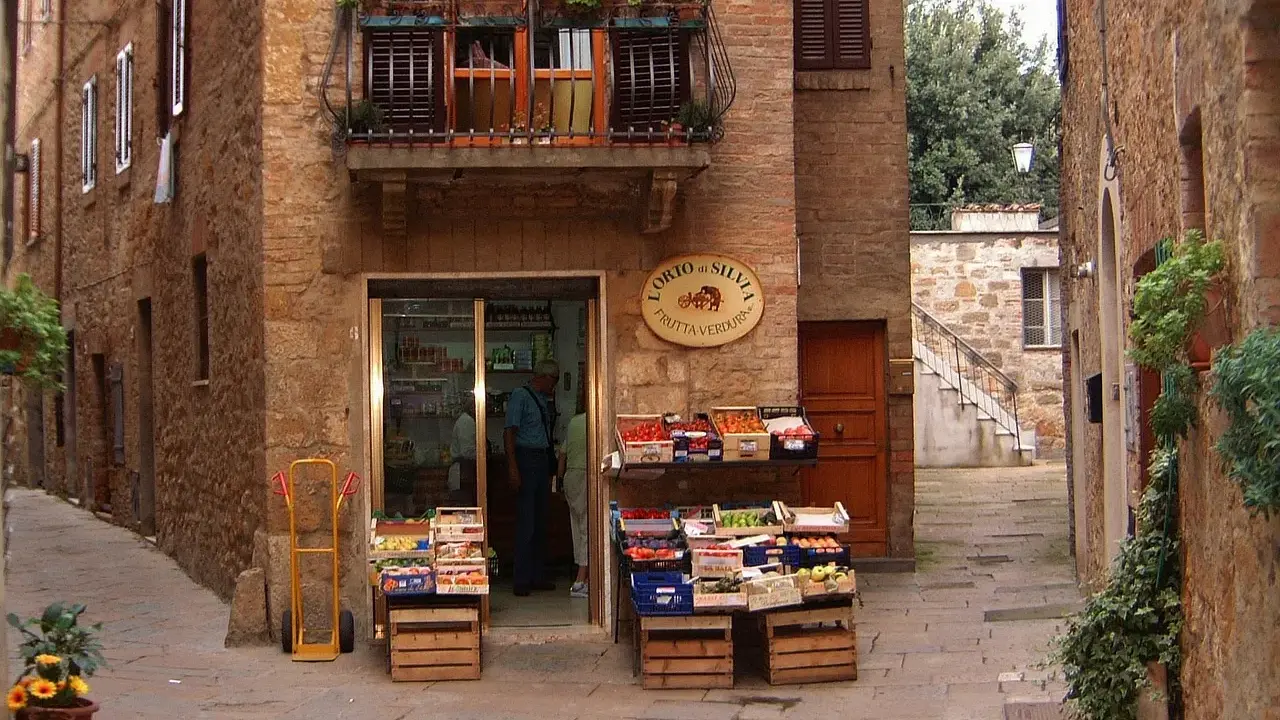Un piccolo negozio di frutta e verdura in un vicolo di **paesi toscani**. Frutta fresca in cassette di legno, un balcone fiorito e l'atmosfera autentica.