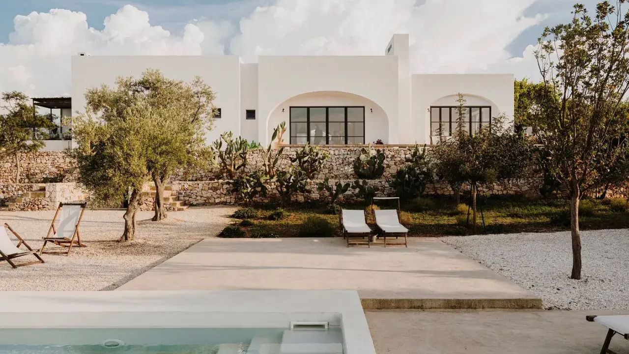 Piscina e lettini in una delle masserie famose puglia, con ulivi e cactus sotto un cielo azzurro con nuvole bianche.