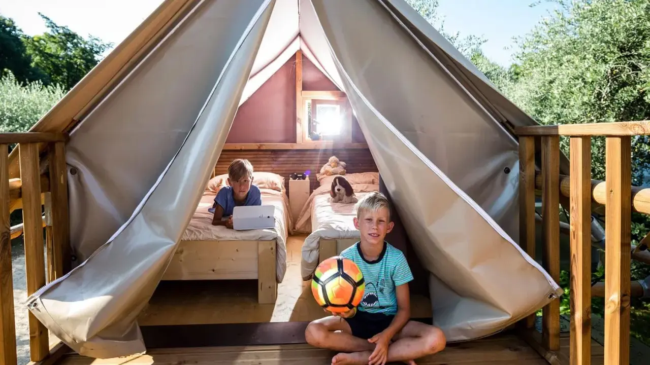 Bambini felici in un glamping: uno gioca col pallone, l'altro usa un tablet a letto. Avventura e relax per i più piccoli.