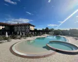 Piscina a forma di rene con idromassaggio annesso, circondata da lettini e ombrelloni, di fronte a un hotel in stile piemontese.