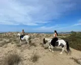 Due donne godono di una passeggiata a cavallo in Sicilia, tra dune sabbiose e il mare azzurro.