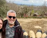 Tre amici sorridono mentre salutano i maiali senesi in un recinto fangoso, con un paesaggio collinare sullo sfondo.