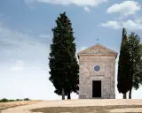 La cappella della Madonna di Vitaleta, con i suoi cipressi iconici, si erge solitaria su una collina toscana sotto un cielo azzurro e nuvoloso.
