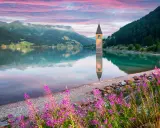 Torre sommersa in un lago alpino, con montagne verdi e cielo rosa all'alba. Un angolo di montagna Friuli Venezia Giulia incantevole.