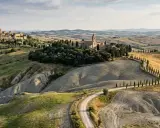 Dolci colline senesi con un borgo medievale e un'abbazia, attraversate da una strada sinuosa fiancheggiata da cipressi.