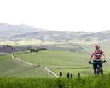 Ciclista in un paesaggio collinare della Toscana, con un sentiero tortuoso che serpeggia tra campi verdi.