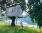 Bambina corre verso una casa sull'albero in legno, con un lago e montagne sullo sfondo.