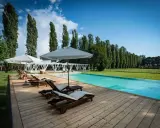 Una splendida piscina in campagna, con lettini prendisole in legno e ombrelloni bianchi, circondata da un prato verde e alti alberi.