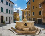 Fontana in pietra nella Valdichiana Senese, circondata da edifici storici con persiane verdi e un cielo azzurro con nuvole bianche.