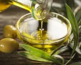 Olio di oliva non extravergine versato in una ciotola di vetro, con olive e foglie d'ulivo su fondo di legno.