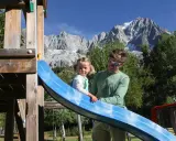Un padre e una bambina si divertono su uno scivolo in un parco giochi con le maestose montagne della Valle d'Aosta sullo sfondo, un'estate indimenticabile.