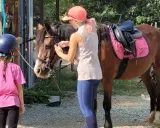 Bambina con casco blu e maglietta rosa osserva un cavallo marrone pronto per un giro a cavallo per bambini. Una ragazza con cappellino arancione prepara il cavallo.