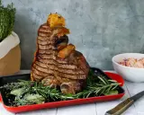 Una succulenta bistecca fiorentina, grigliata alla perfezione, adagiata su erbe aromatiche in una teglia rossa. Accanto, un'insalata fresca e un coltello.