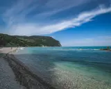 Spiaggia di ciottoli con mare cristallino e una collina verdeggiante, tipica del entroterra marchigiano.