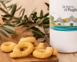 Taralli pugliesi su tagliere di legno, accanto a un contenitore con il logo "la terra di Puglia" e un ramo d'ulivo.