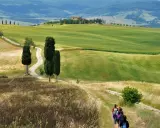 Un gruppo di escursionisti vive le **esperienze Val d'Orcia** su un sentiero tra colline verdi e cipressi, con una casa colonica in lontananza.