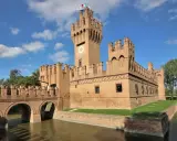 Un imponente castello in mattoni rossi, uno dei tanti castelli Emilia Romagna, con un ponte ad archi sull'acqua e torri merlate sotto un cielo azzurro.