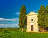 Cappella di San Quirico d'Orcia, circondata da cipressi, su un prato verde sotto un cielo azzurro.